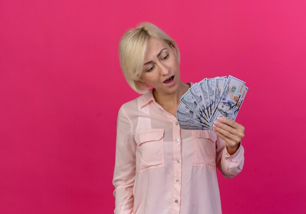 Impressed young blonde slavic woman holding and looking at money isolated on pink background with copy space