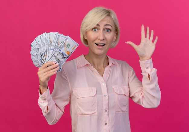 Impressed young blonde slavic woman holding money and gesturing hi with hand isolated on pink background