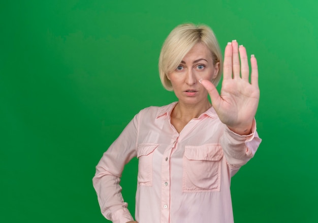 Впечатленная молодая белокурая славянская женщина смотрит в камеру и делает стоп-жест, изолированные на зеленом фоне с копией пространства