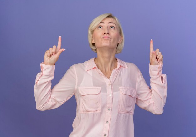 Impressed young blonde slavic woman looking and pointing up isolated on purple background