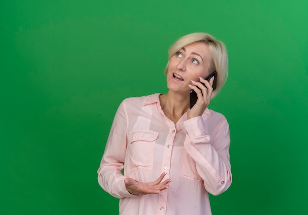 Impressionato giovane bionda donna slava alzando lo sguardo parlando al telefono tenendo la mano in aria isolato su sfondo verde con copia spazio