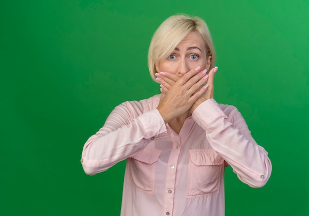 Impressed young blonde slavic woman putting hands on mouth isolated on green background with copy space