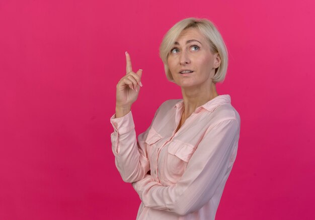 Impressed young blonde slavic woman standing in profile view looking and pointing up isolated on pink background with copy space