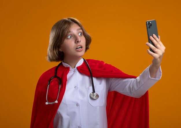 Impressed young blonde superhero woman in red cape wearing doctor uniform and stethoscope taking selfie isolated on orange wall