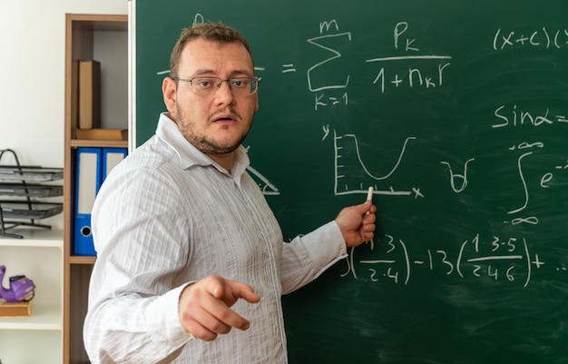 Impressed young blonde teacher wearing glasses standing in profile view in front of chalkboard in classroom looking and pointing at camera pointing at chalkboard with chalk