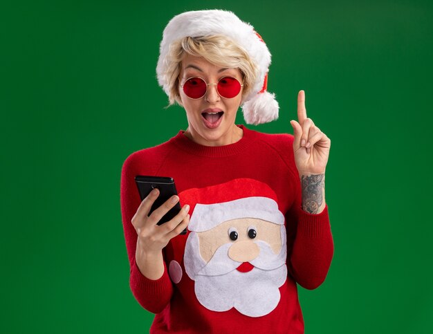 크리스마스 모자와 산타 클로스 크리스마스 스웨터를 입고 감동 된 젊은 금발의 여자 안경 들고와 복사 공간이 녹색 벽에 고립 가리키는 휴대 전화를 찾고
