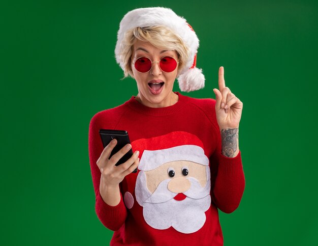 Impressed young blonde woman wearing christmas hat and santa claus christmas sweater with glasses holding and looking at mobile phone pointing up isolated on green wall with copy space