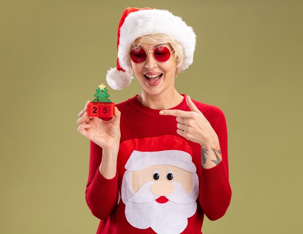크리스마스 모자와 산타 클로스 크리스마스 스웨터를 입고 감동 된 젊은 금발의 여자 안경을보고 및 복사 공간 올리브 녹색 벽에 고립 된 날짜와 함께 크리스마스 트리 장난감을 가리키는