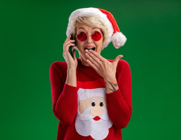 Foto gratuita impressionato giovane donna bionda che indossa il cappello di natale e babbo natale maglione di natale con gli occhiali parlando al telefono guardando la telecamera tenendo la mano sulla bocca isolata su sfondo verde