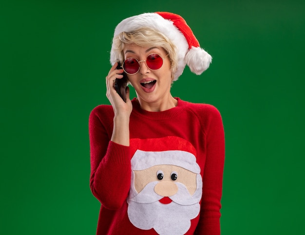 크리스마스 모자와 산타 클로스 크리스마스 스웨터를 입고 감동 된 젊은 금발의 여자 안경 녹색 배경에 고립 내려다보고 전화 통화