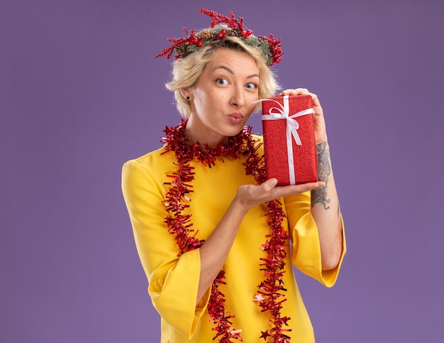 クリスマスの頭の花輪と首の周りに見掛け倒しの花輪を身に着けている印象的な若いブロンドの女性は、コピースペースで紫色の壁に分離されたすぼめた唇で見ているギフトパッケージを保持しています