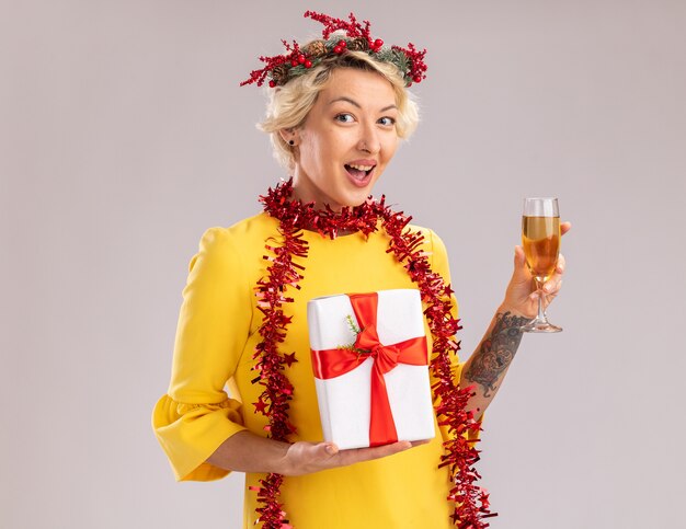 Impressed young blonde woman wearing christmas head wreath and tinsel garland around neck holding glass of champagne and gift package looking at camera isolated on white background