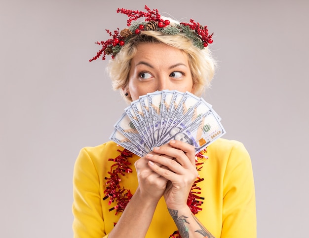 Впечатленная молодая блондинка в рождественском венке и гирлянде из мишуры на шее, держащая деньги, смотрящую в сторону сзади, изолированную на белом фоне