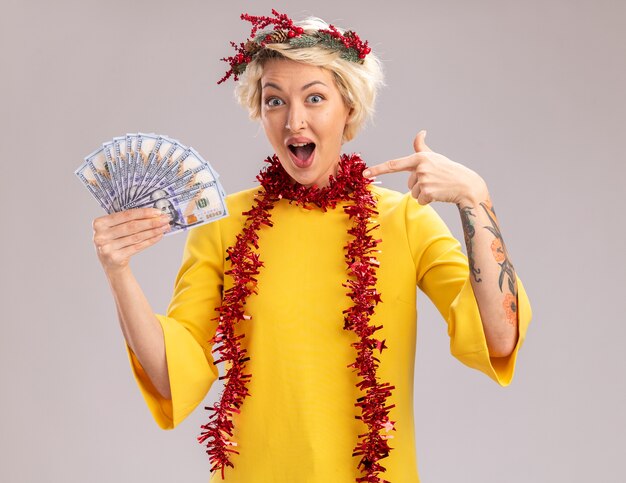 Impressed young blonde woman wearing christmas head wreath and tinsel garland around neck holding and pointing at money looking  isolated on white wall