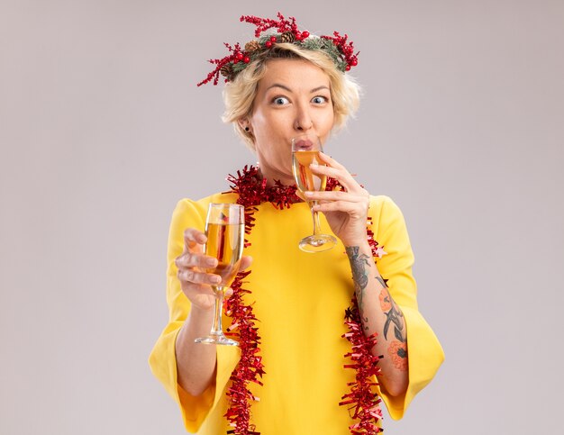 Впечатленная молодая блондинка в рождественском венке и гирлянде из мишуры на шее, держащая два бокала шампанского, глядя в камеру, пьет один на белом фоне