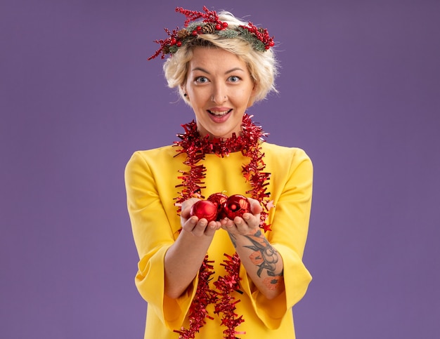 Impressionato giovane donna bionda che indossa la testa di natale ghirlanda e orpello ghirlanda intorno al collo guardando la telecamera tenendo le bagattelle di natale isolato su sfondo viola