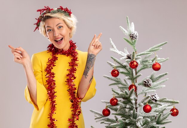 Impressed young blonde woman wearing christmas head wreath and tinsel garland around neck standing near decorated christmas tree looking at camera pointing up isolated on white background