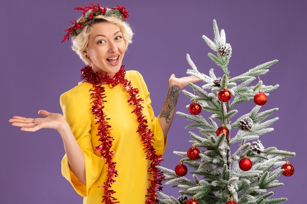 Впечатленная молодая блондинка в рождественском венке и гирлянде из мишуры на шее, стоящая возле украшенной елки, глядя в камеру, показывая пустые руки, изолированные на фиолетовом фоне