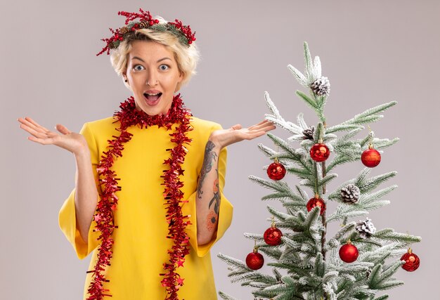 Impressed young blonde woman wearing christmas head wreath and tinsel garland around neck standing near decorated christmas tree looking at camera showing empty hands isolated on white background