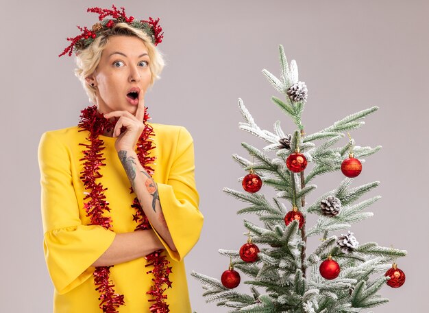 Impressed young blonde woman wearing christmas head wreath and tinsel garland around neck standing near decorated christmas tree looking at camera touching lips isolated on white background