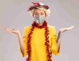 Free photo impressed young blonde woman wearing christmas head wreath and tinsel garland around neck with protective mask looking at camera showing empty hands isolated on white background