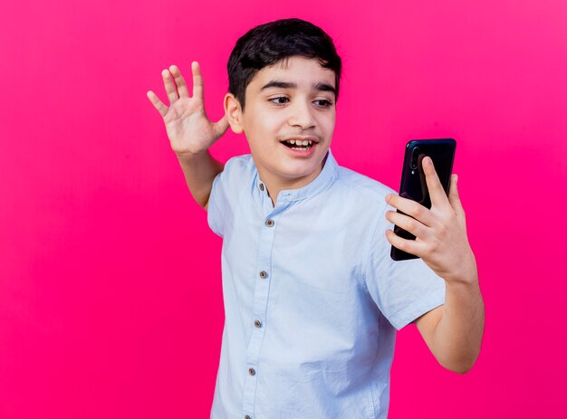 Impressed young boy holding and looking at mobile phone keeping hand in air isolated on pink wall