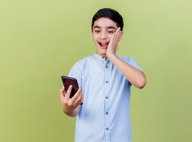 Impressed young boy holding and looking at mobile phone keeping hand on face isolated on olive green wall