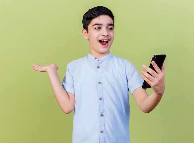 Impressed young boy holding and looking at mobile phone showing empty hand isolated on olive green wall