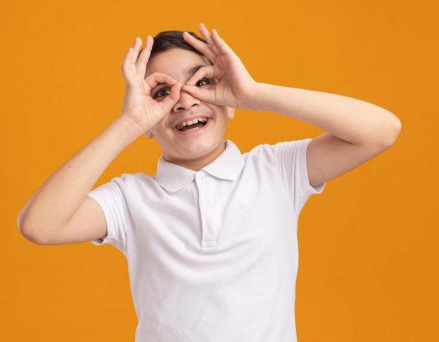 Impressed young boy looking at front doing look gesture using hands as binoculars isolated on orange wall