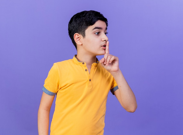 Impressed young boy looking at side doing silence gesture isolated on purple wall