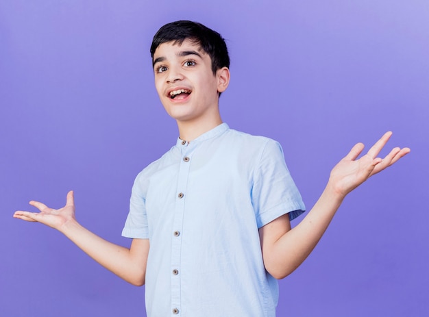 Impressed young boy looking at side showing empty hands isolated on purple wall