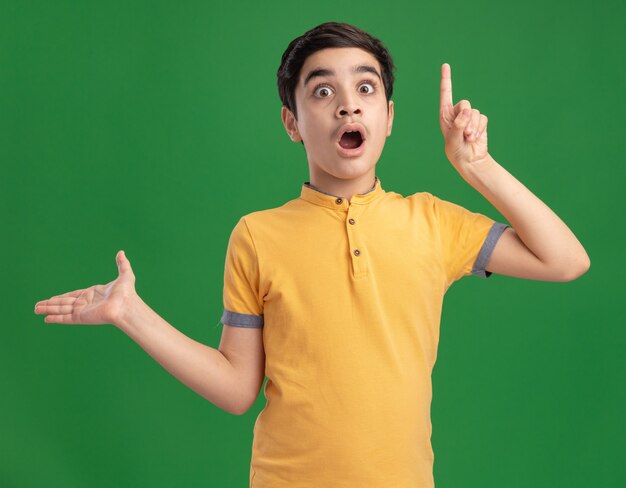 Impressed young boy pointing up showing empty hand looking at front isolated on green wall