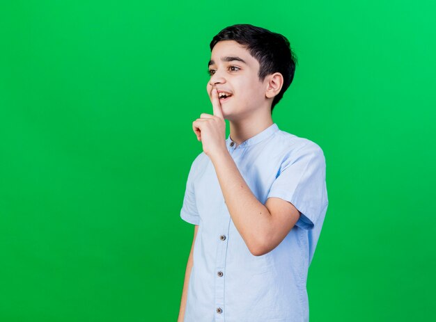 Impressed young boy standing in profile view looking straight doing silence gesture isolated on green wall