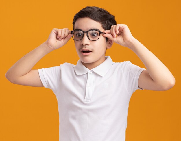 Impressed young boy wearing and grabbing glasses looking down isolated on orange wall