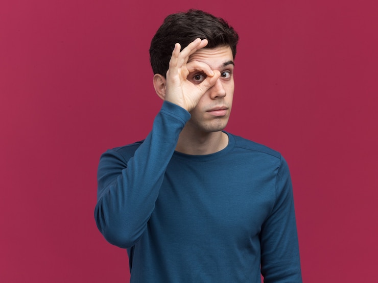 Free Photo impressed young brunette caucasian boy looking at camera through fingers on pink