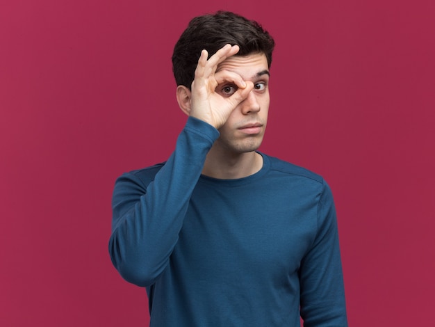 Impressed young brunette caucasian boy looking at camera through fingers on pink