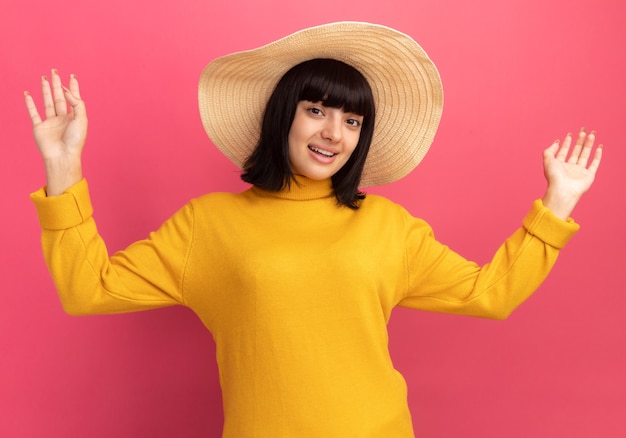 Impressed young brunette caucasian girl wearing beach hat stands with raised hands on pink 