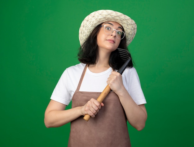 Impressed young brunette female gardener in optical glasses and uniform wearing gardening hat holds rake isolated on green wall