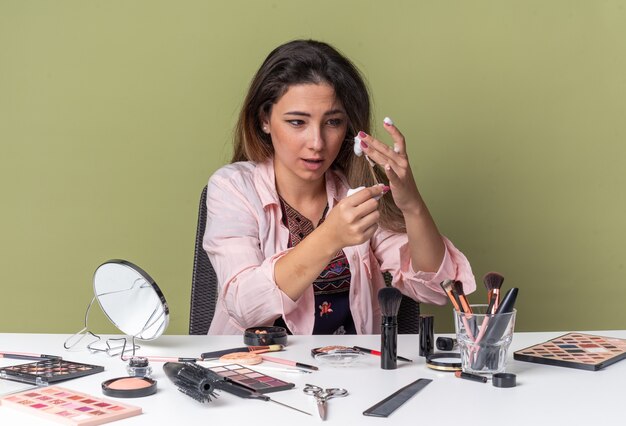 Impressed young brunette girl sitting at table with makeup tools applying hair mousse