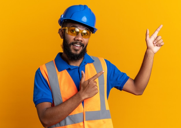 impressed young builder man in safety glasses wearing uniform with safety helmet pointing at side isolated on orange wall with copy space