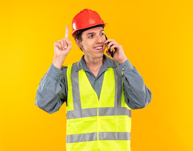 Impressed young builder man in uniform speaks on phone points at up isolated on yellow wall