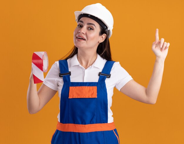 Impressed young builder woman in uniform holding duct tape points at up isolated on orange wall