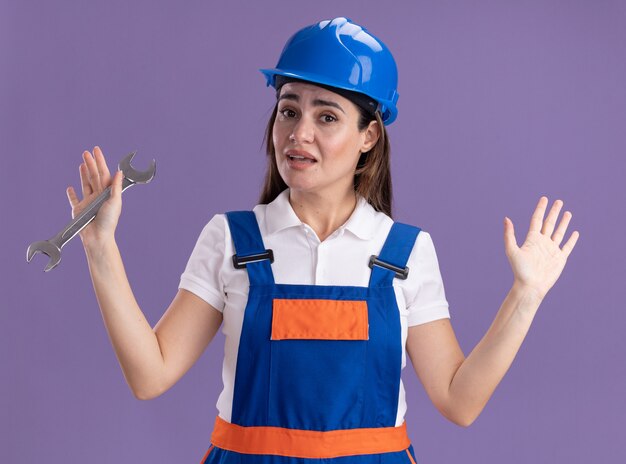 보라색 벽에 고립 된 손을 확산 오픈 엔드 렌치를 들고 유니폼에 감동 된 젊은 작성기 여자