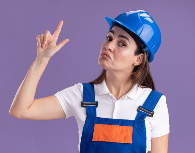 Впечатленная молодая женщина-строитель в униформе указывает вверх, изолированную на фиолетовой стене