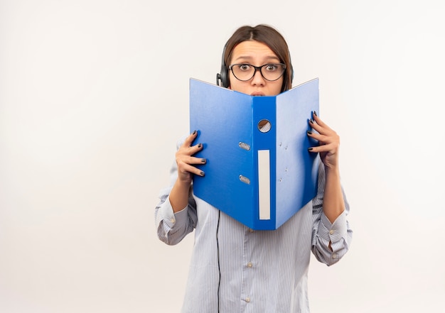 Free photo impressed young call center girl wearing glasses and headset holding and looking from behind folder isolated on white  with copy space