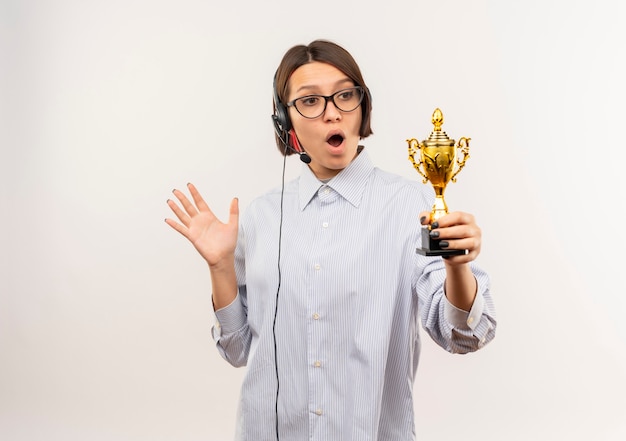 Impressed young call center girl wearing glasses and headset holding and looking at winner cup and showing empty hand isolated on white  with copy space