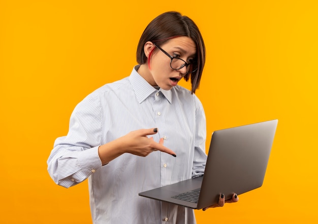 Impressed young call center girl wearing glasses holding looking and pointing at laptop isolated on orange 