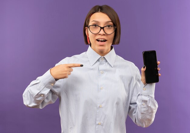Impressed young call center girl wearing glasses holding and pointing at mobile phone isolated on purple 