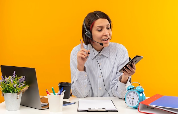 Impressionato giovane ragazza della call center che indossa la cuffia avricolare che si siede allo scrittorio che tiene e che esamina il telefono cellulare con il dito alzato isolato sull'arancio