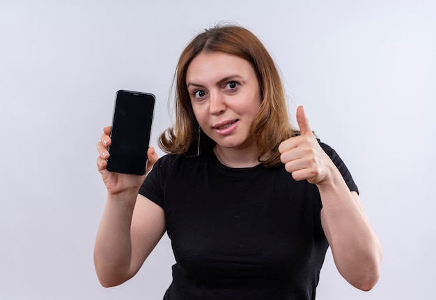 Impressed young casual woman holding mobile phone and showing thumb up on isolated white space